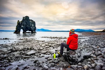 A visitor stands on a black sand beach, looking at Hvitserkur Rock, an iconic basalt formation in Iceland. The sky is overcast, creating a dramatic scene. © Kotangens