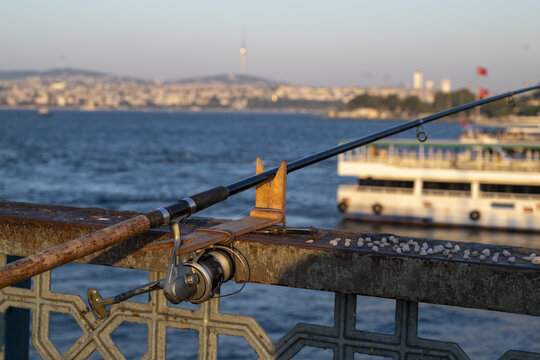 View of a fishing rod resting on a bridge railing overlooking the Bosphorus with a ferry and the city skyline in the background in Istanbul, Istanbul, Turkiye.
