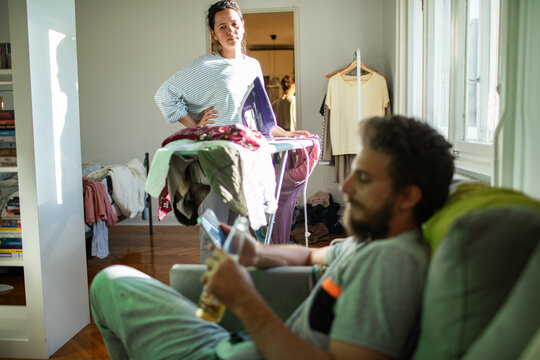 Frustrated woman ironing while partner relaxes on sofa at home