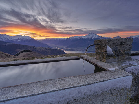 View of a stone water trough with flowing water set against a dramatic sunset over the snow-capped mountains and Lake Como in Gera Lario, Lombardia, Italy.