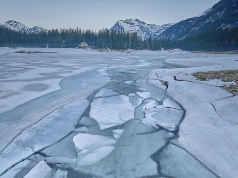 View of the frozen Lago Palu with cracked ice sheets on the surface, surrounded by snow-covered pine forests and rugged mountain peaks in Chiesa in Valmalenco, Lombardia, Italy.