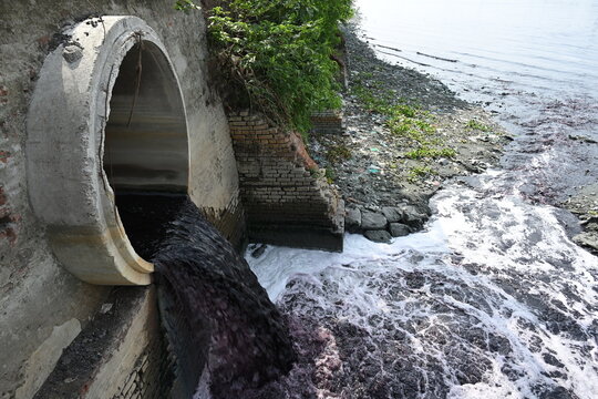 Dhaka, Bangladesh - 14 September 2026: View of a concrete drainage pipe discharging dark, polluted wastewater into a river, with foam and debris near the brick embankment.