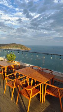 Empty wooden table and chairs setup on a rooftop cafe balcony overlooking a calm blue sea, coastal mountain landscape, and green tropical plants under a partly cloudy sky. Perfect background for