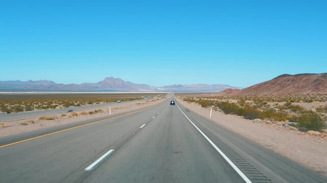 Car driver point of view driving on straight endless road through arid landscape of mojave desert in california, with distant mountains and clear blue sky on the horizon. Road trip on modern highway