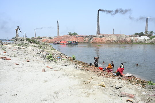 Dhaka, Dhaka Division, Bangladesh - 14 September 2026: View of people washing clothes in the river near brick kilns with smoking chimneys in the background.