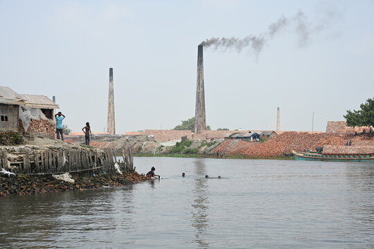 Dhaka, Bangladesh - 14 September 2026: View of brick kilns with tall chimneys emitting smoke, piles of bricks, and people swimming in the river in Dhaka.
