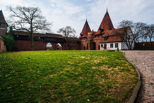 Malbork, Poland - 23 March 2026: View of Malbork Castle, featuring its iconic red brick fortifications, tall towers, and a grassy courtyard under a cloudy sky.