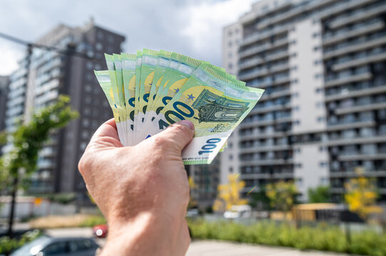 Male Hand Holding Stack of 100 Euro Banknotes Against Modern Apartment Buildings Exterior