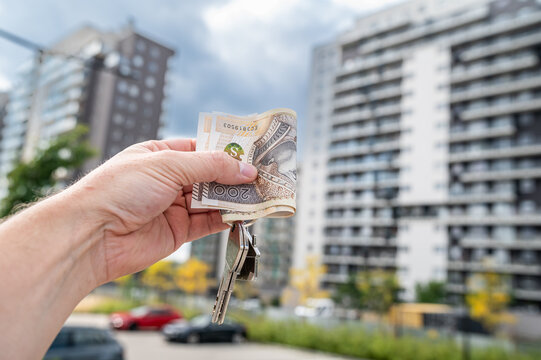 Male Hand Holding House Keys with 200 Polish Zloty Banknotes Against Modern Apartment Building