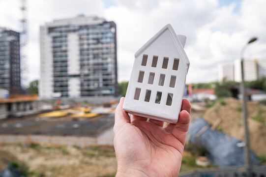 Male Hand Holding White House Model in Front of Modern Apartment Building Construction Site