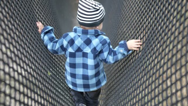 A child in a checked jacket moves along a flexible net bridge while gripping its sides, showing a playground obstacle used for balance and physical play