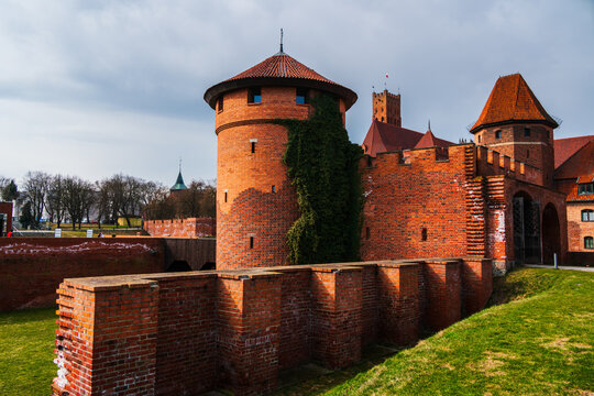 Malbork, Poland - 23 March 2026: View of Malbork Castle with its iconic red brick defensive walls, towers, and ivy-covered masonry under a cloudy sky.