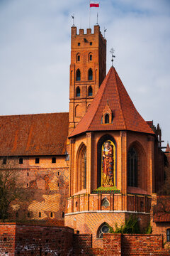 Malbork, Poland - 23 March 2026: View of the historic Malbork Castle, featuring iconic red brick architecture, a tall tower with a Polish flag, and a statue of the Virgin Mary.