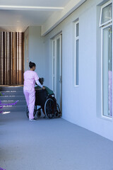 Naklejka premium African American caregiver in pink scrubs pushing Asian senior in wheelchair at door, copy space