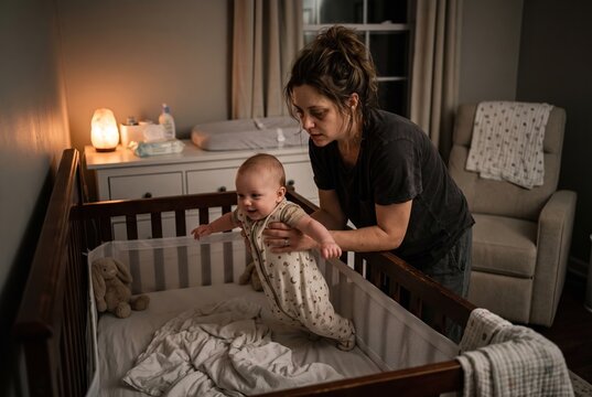 A mother assists her baby in standing inside a crib in a nursery. The room is softly lit, creating a cozy setting. The baby appears eager and playful