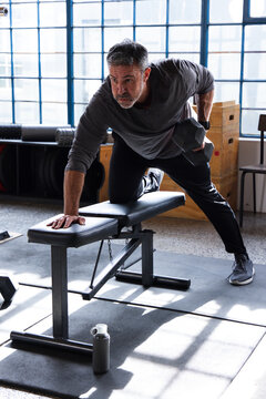Middle-aged man in gray top performing bent-over row pulling hex dumbbell bracing on bench in gym