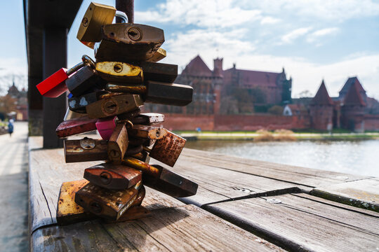 Malbork, Poland - 23 March 2026: View of love padlocks attached to a wooden bridge railing with the historic Malbork Castle in the background under a blue sky.