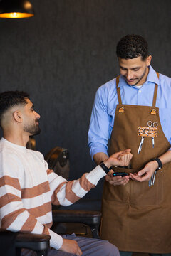 African male barber in apron holding reader while male customer holding smartphone at barbershop