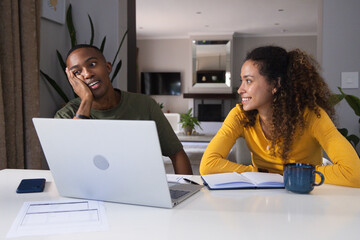Obraz premium Couple sitting at open-plan living room table, sorting papers with laptop, mustard top, olive tee