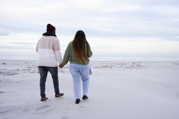 Obraz premium African couple walking hand in hand on sandy beach to shoreline, wearing beanie, leaving footprints