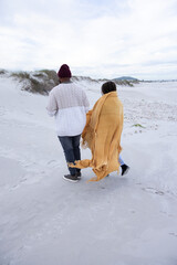 Obraz premium Couple walking across pale sand dunes at beach, showing mustard blanket, beanie, sweater