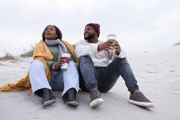 Obraz premium African American couple sitting on sandy dune at beach holding reusable travel cups, wearing shawl