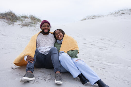 African American couple sitting on sandy beach dune sharing mustard blanket, smiling
