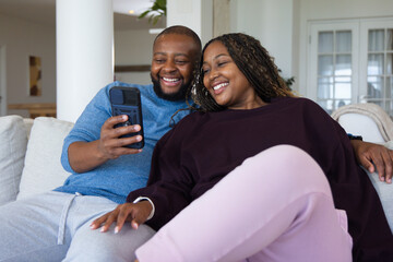 Obraz premium African American couple sitting on light sofa in living room sharing smartphone with bracelet