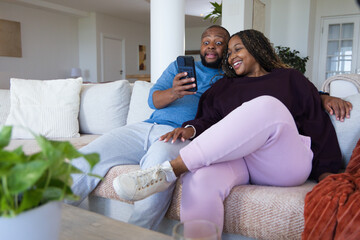 Obraz premium African couple sitting on light sectional sofa at home, man holding smartphone, woman smiling