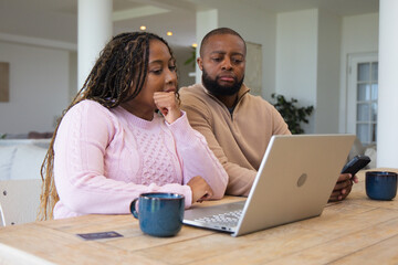 Obraz premium Couple sitting at wooden table at home, reviewing laptop with smartphone and woman in pink sweater
