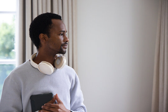 Adult man wearing sweater and headphones standing at home by window holding tablet, facing right