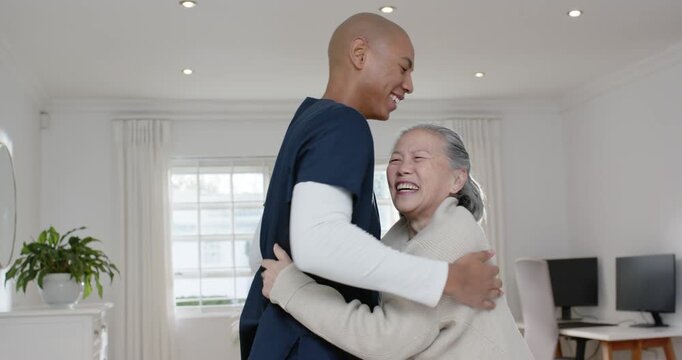 Senior Asian woman, African American in navy scrubs visiting home hugging smiling opening cardigan