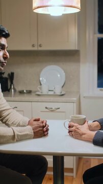 Vertical video: Conversation starting, couple in sweaters leaning at kitchen table, holding mug