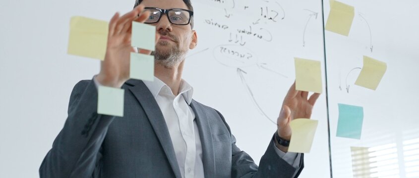 Businessman in glasses organizing sticky notes on a transparent glass wall, planning a corporate strategy and outlining key points for a new business project