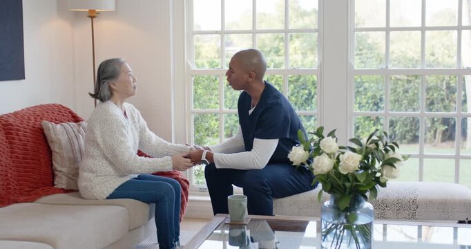 Senior Asian woman on sofa, caregiver scrubs reaching, holding hands, reassuring by window with mug