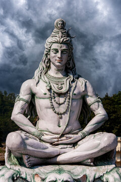 Lord Shiva statue meditating against dramatic stormy sky. Majestic white stone sculpture of Hindu god Shiva sitting lotus pose and meditating under storm clouds, India, travel and religion concept
