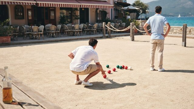 Two men playing a game of bocce on a sandy court near a seaside restaurant with outdoor court located bocce colorful balls recreational activity