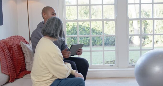 African American man tapping tablet guiding Senior Chinese woman to ball for rehab in living room