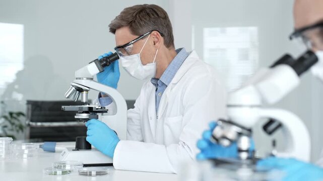 Researcher in a lab coat and protective gear is performing microbiology work, looking through a microscope to analyze scientific samples for medical research and development