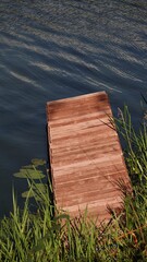 Drewniany pomost wysunięty w głąb spokojnej wody, otoczony zieloną trawą i liśćmi lilii wodnych. Spokojny, naturalny kadr przedstawiający urokliwy zakątek nad jeziorem © tomo81