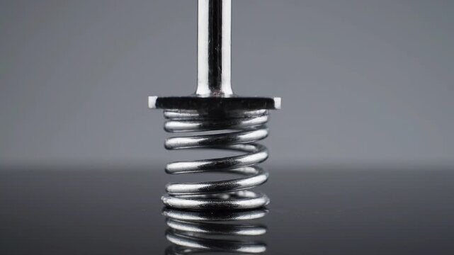 Spring loaded metal plunger moves up and down with precise industrial motion. Metal spring assembly appears metallic, reflective, and focused on repeated compression against gray backdrop.