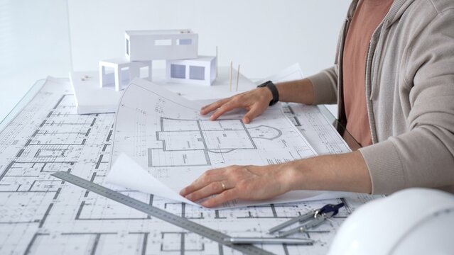 Professional architect's hands study detailed blueprints and building plans on a drafting table, with a 3d house model nearby, focusing on residential design and engineering decisions