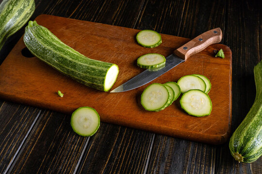 A vibrant green zucchini is being sliced on a dark wooden cutting board. The knife glistens as it cuts through the tender flesh, creating perfect rounds