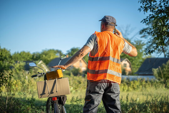 A delivery worker in an orange vest stands beside his bicycle, making a phone call against a backdrop of green trees and houses. The sun shines brightly on this busy day
