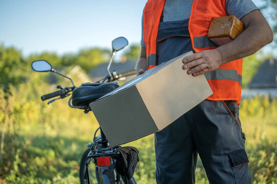 A delivery courier stands beside a bicycle in a sunny outdoor setting, grasping a package ready for delivery. Trees and a clear sky frame the scene, highlighting a busy day ahead
