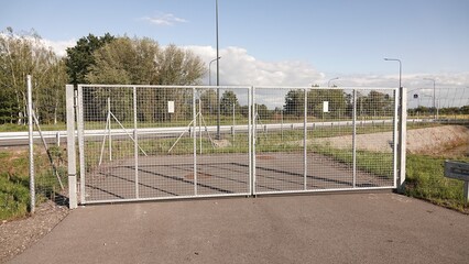 Zamknięta, dwuskrzydłowa brama panelowa zabezpieczająca wjazd na asfaltową drogę. W tle widoczna infrastruktura drogowa, zieleń oraz błękitne niebo z chmurami. © tomo81