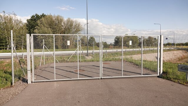 Zamknięta, dwuskrzydłowa brama panelowa zabezpieczająca wjazd na asfaltową drogę. W tle widoczna infrastruktura drogowa, zieleń oraz błękitne niebo z chmurami.