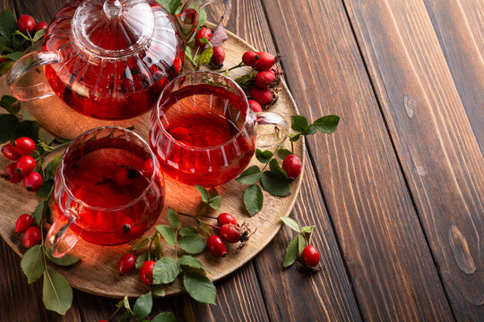 Glass teapot and cups filled with red rosehip tea next to fresh berries and green leaves on wooden tray