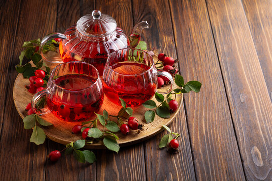 Rosehip tea brewed in glass teapot served in cups with fresh rosehip berries on wooden tray