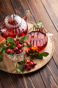 Rosehip tea in glass teapot and cup served on wooden tray with fresh berries and leaves